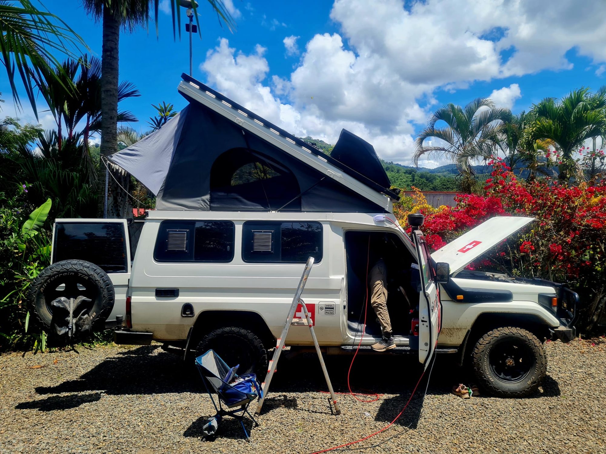 Auto mit geöffneten Dachzelt und Hube. Mann zieht ein neues Kabel von den Solarpanels zur Batterie.